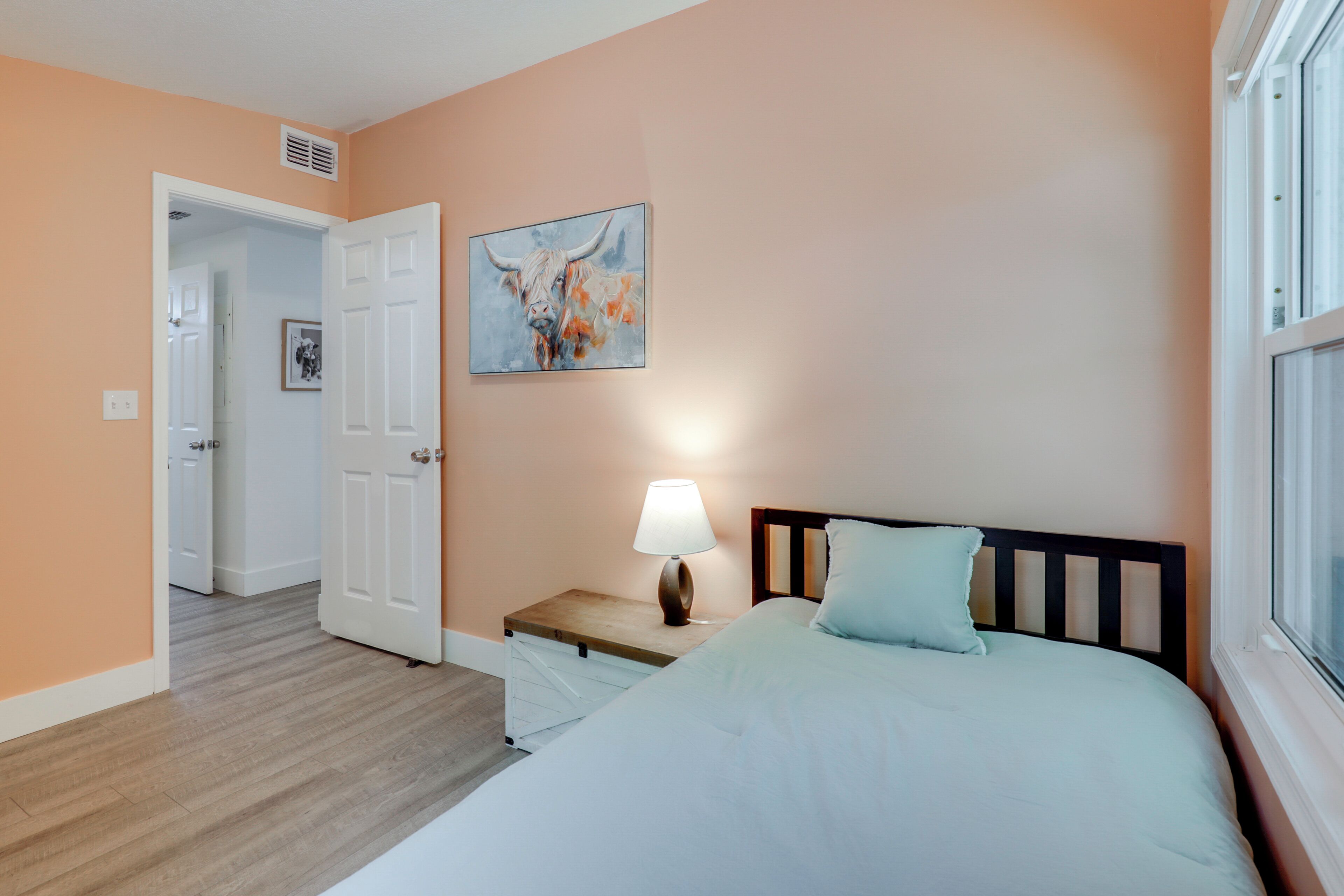 A bedroom with a bed, a nightstand with a lamp, a painting on the wall, and a door leading to another room.