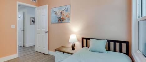A bedroom with a bed, a nightstand with a lamp, a painting on the wall, and a door leading to another room.