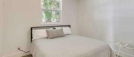 Un dormitorio con cama, ventana y pared blanca.