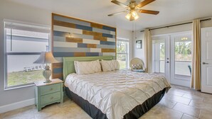 Quarto com cama, mesa de cabeceira, ventilador de teto, janela com persianas e porta.