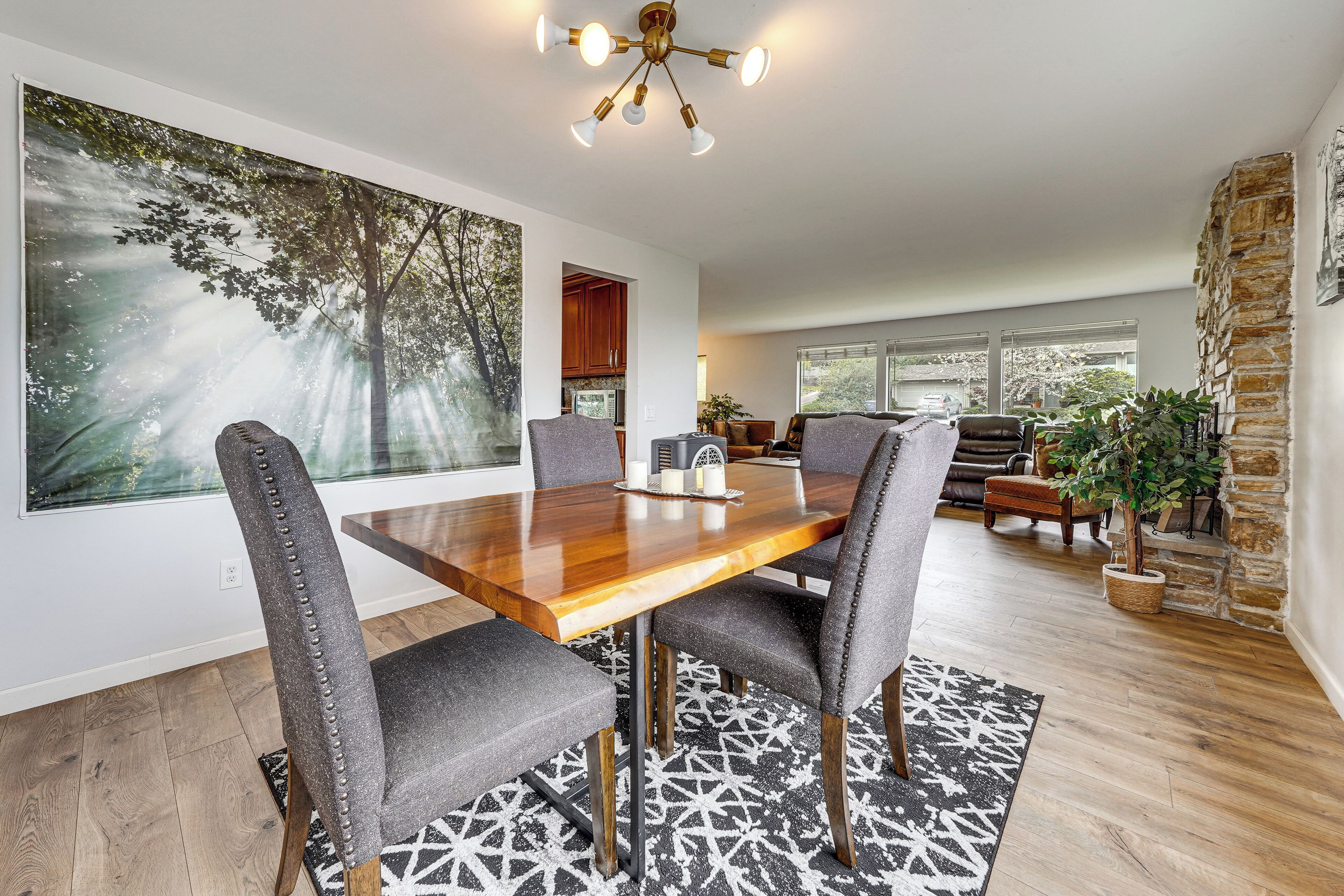 Uma sala de jantar com mesa de madeira, cadeiras estofadas cinzentas, um quadro grande com tema de floresta e uma parede de pedra decorativa.
