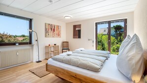 Ein modernes Schlafzimmer mit einem Holzbett, einem Heizkörper, einer Stehlampe, einem Sessel und Blick auf Grünfläche.