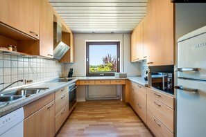 A modern kitchen with wooden cabinets, a built-in oven, and a window with a view of greenery. - Vacation apartment Aussichtsreich - spacious with terrace and outdoor fireplace (Lindau (Bodensee))