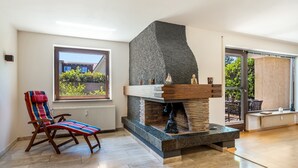 Ein modernes Wohnzimmer mit Kamin aus Stein, einem farbenfrohen Sessel und Blick durch ein großes Fenster.