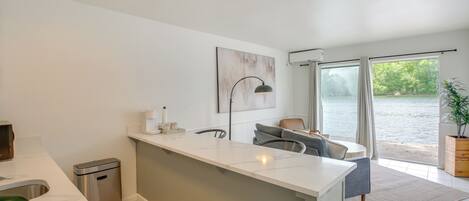 A modern kitchen with a white countertop, a trash bin, a gray sofa, and a window overlooking a body of water.