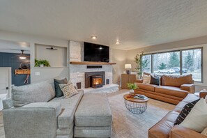 Ein modernes Wohnzimmer mit Kamin aus Stein, grauen und braunen Sofas, einem runden Couchtisch und großen Fenstern.