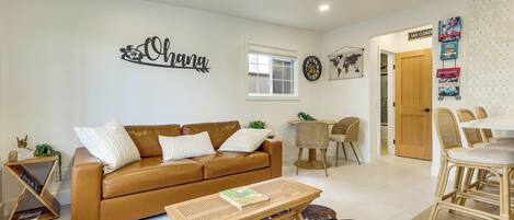 Un salon avec un canapé en cuir marron, une table basse, un coin repas avec des chaises en osier et un mur orné d’une inscription « Ohana ».