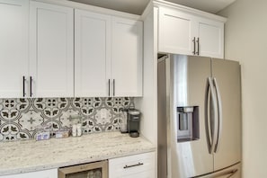 Cocina con gabinetes blancos, refrigerador de acero inoxidable, cafetera y azulejos decorativos en la pared.