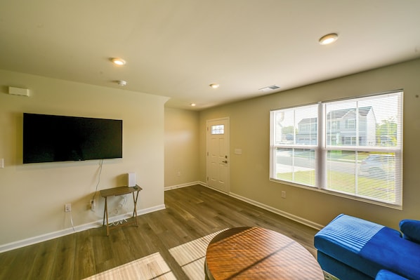 A room with a flat-screen TV mounted on the wall, a round wooden table, a blue sofa, and large windows with blinds.