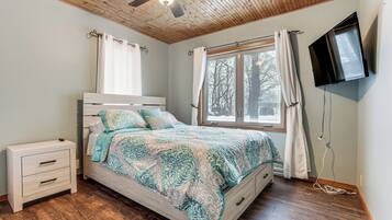 A bedroom with a bed, a nightstand, a ceiling fan, a window with curtains, and a TV mounted on the wall.