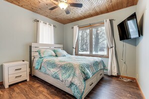 Ein Schlafzimmer mit Bett, Nachttisch, Deckenventilator, Fenster mit Vorhängen und einem an der Wand montierten Fernseher.