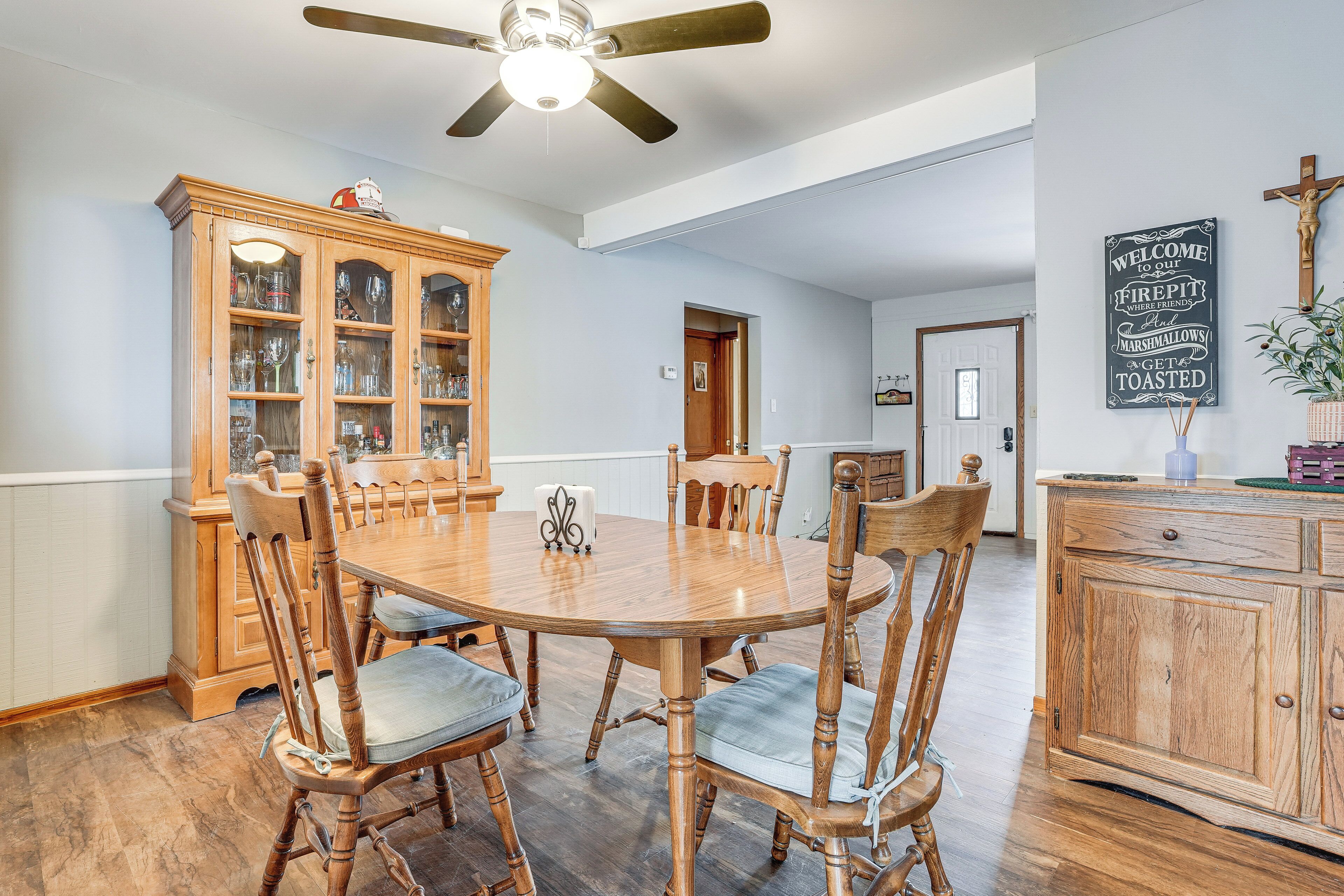A dining room with a wooden table and chairs, a cabinet with glass doors, a ceiling fan, and a wall with a welcome sign.
