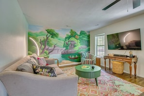 Un salón con un mural, un sofá, una mesa de centro, un televisor y una ventana.