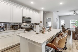 Une cuisine moderne avec des meubles blancs, un grand îlot central doté de tabourets et un coin repas agrémenté d’un ventilateur de plafond.