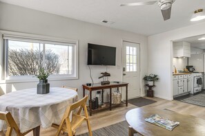 A modern living room with a dining table, chairs, a flat-screen TV, and a kitchen area visible through an open door.