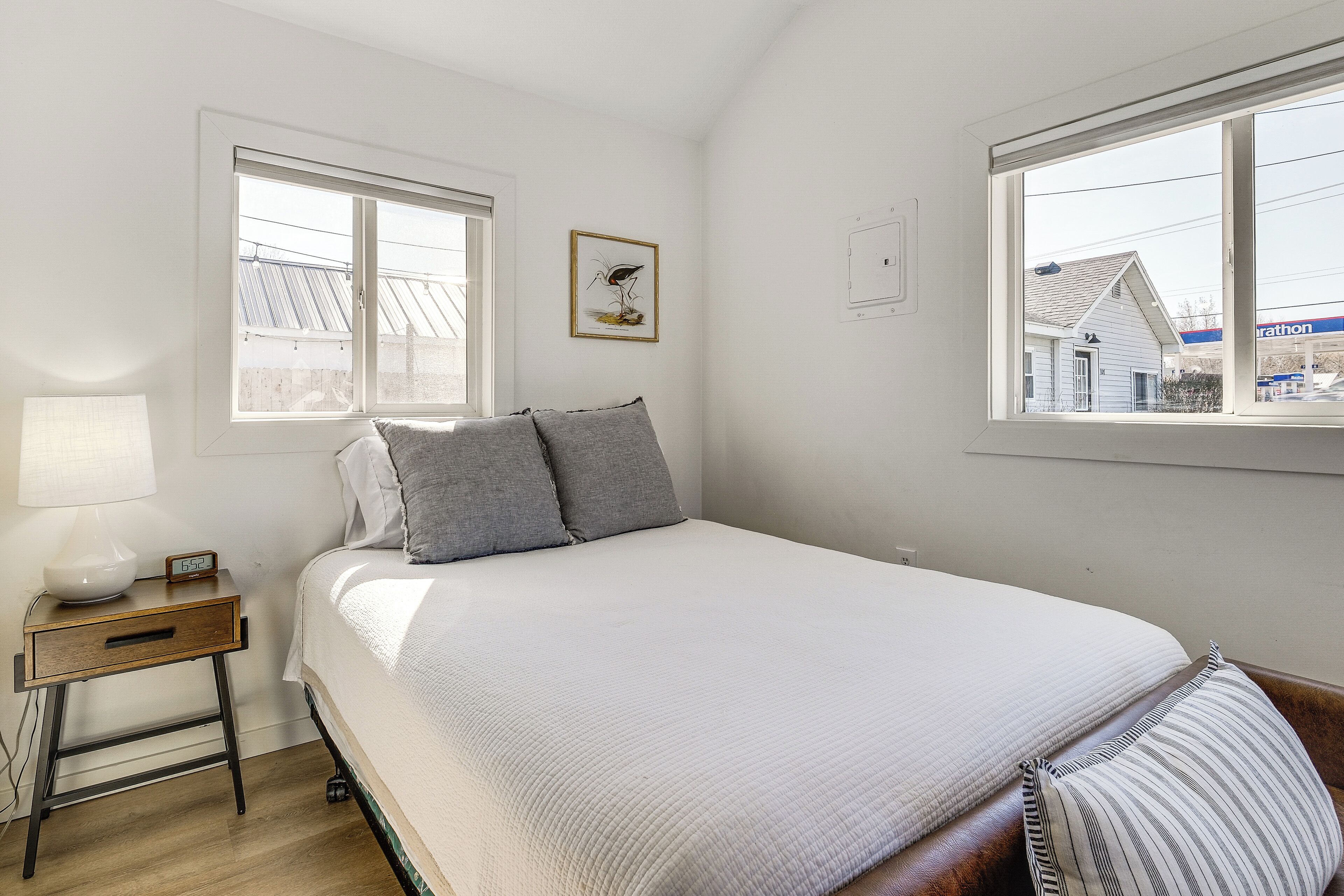 A bedroom with a bed, a nightstand, a lamp, a clock, and a window with a view of a house and a Marathon gas station.