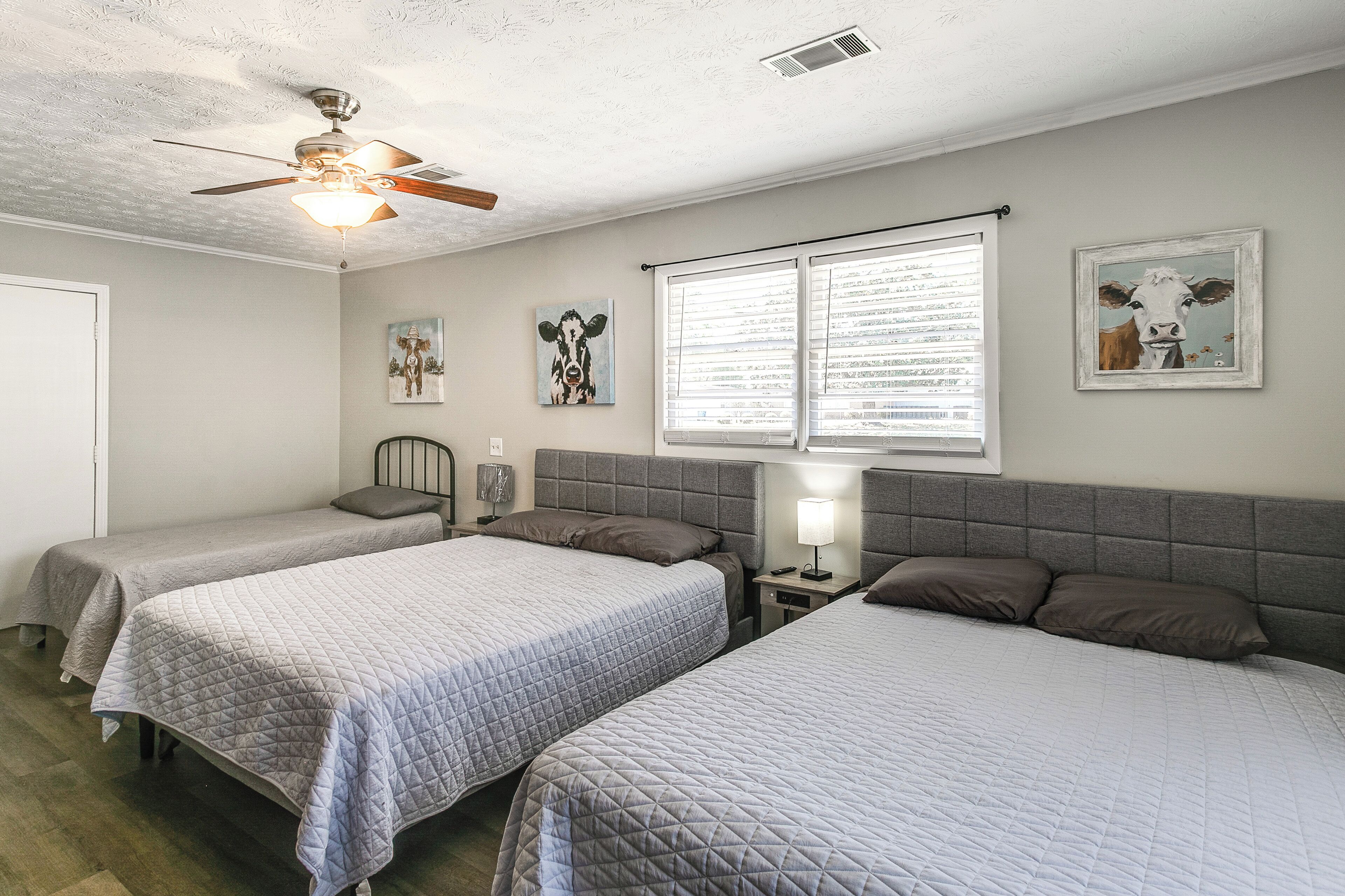 A room with three beds, a ceiling fan, a window with blinds, and wall art.