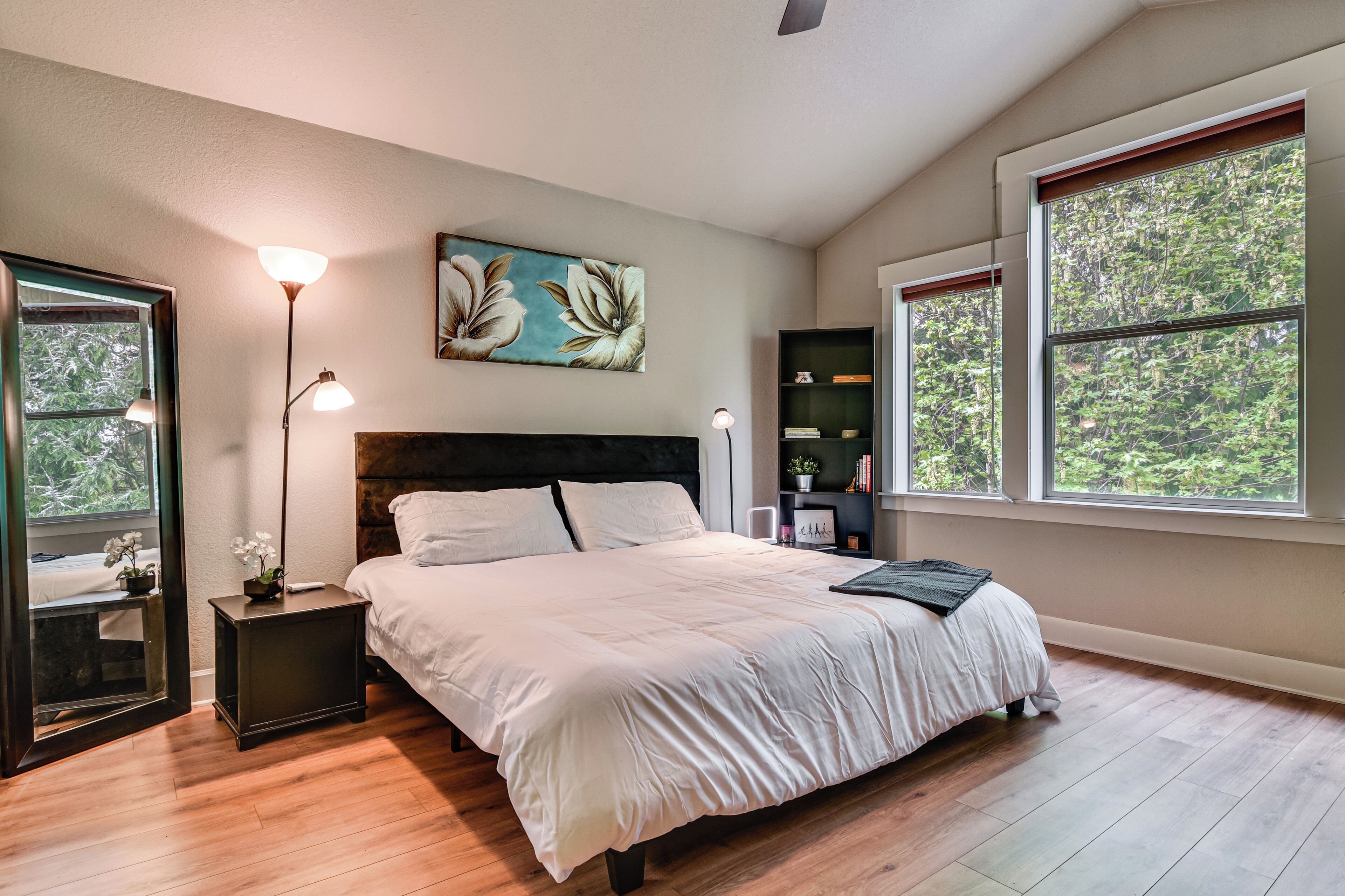 A bedroom with a large bed, bedside tables, a nightstand, a floor lamp, a painting, a bookshelf, and large windows with a view of trees.