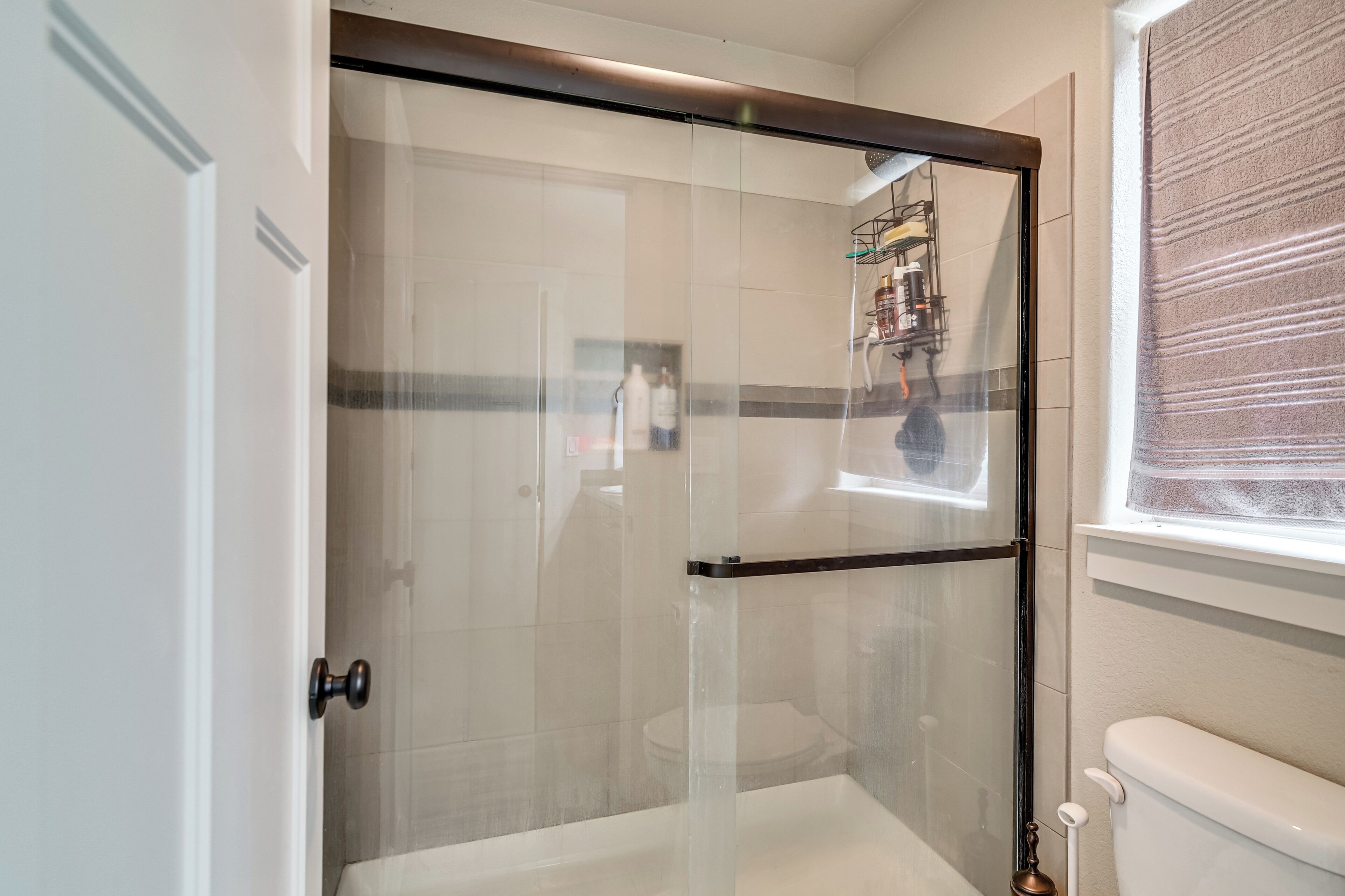 A bathroom with a glass-enclosed shower, a toilet, and a window with blinds.