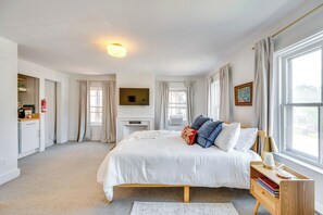 A spacious bedroom with a large bed, a bedside table with books, a flat-screen TV mounted on the wall, and a fireplace. - Walkable 3-unit Retreat on Main Street, Waterbury! (Waterbury)