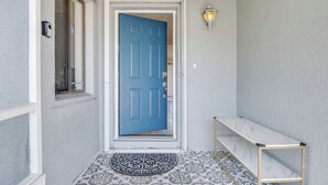 Una puerta azul con marco blanco, un banco pequeño, piso con diseño y una lámpara fijada a la pared.