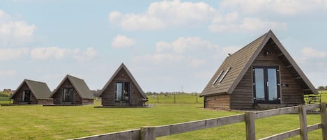 Three wooden triangular cabins with large windows, set in a grassy field with a wooden fence in the foreground.
