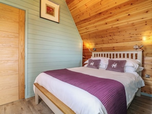 Quarto com teto inclinado, paredes de madeira, cama com roupa de cama branca e roxa, e um quadro pendurado na parede.