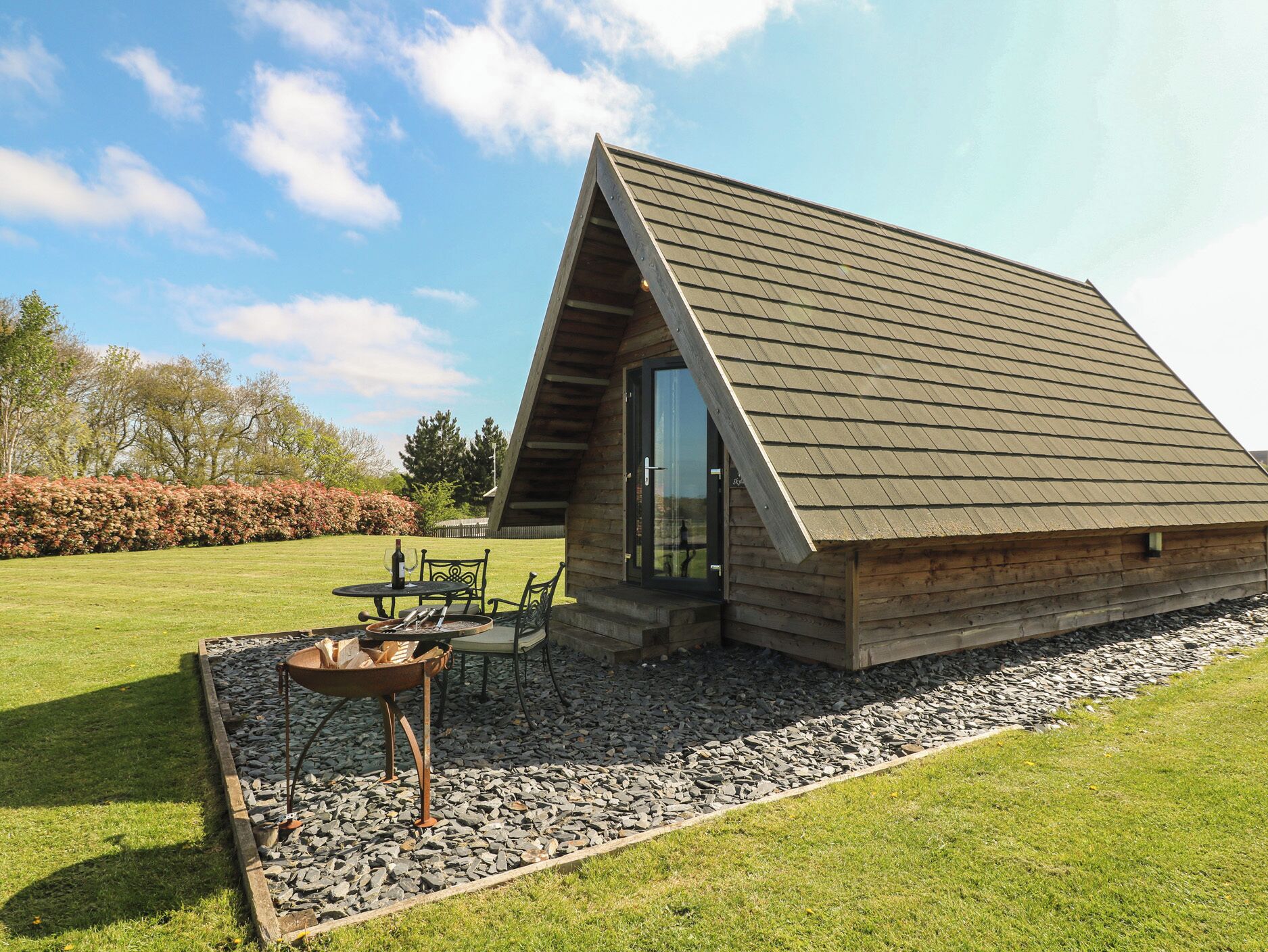 A wooden cabin with a sloped roof, a patio with a table and chairs, and a gravel pathway.