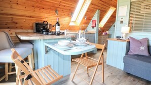 Interior acogedor de una cabaña con techo de madera, un comedor con mesa y sillas, una cocina con hervidor y una botella de vino, y una pared azul de acento.