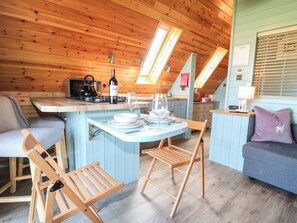 A cozy cabin interior with a wooden ceiling, a dining area with a table and chairs, a kitchen with a kettle and wine bottle, and a blue accent wall. - Skylark (Loughborough)