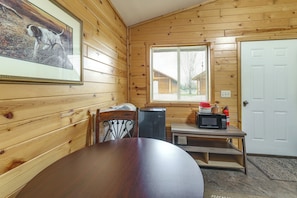 A wooden interior with a dining table, chairs, a framed picture, a window, a door, and a microwave on a small table.
