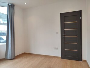 Una habitación con piso de madera, una puerta oscura y una ventana con cortinas grises.