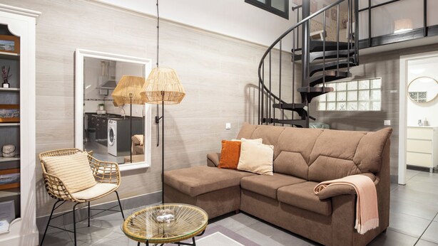 Un salon comprenant un canapé, un fauteuil et une table ronde, un escalier, et une cuisine visible à travers une porte vitrée.