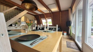 A dining area with a wooden table set for two, a staircase, and a view through large windows. - Storchennest roof-only house: Comfortable vacation home on the North Sea in Butjadingen (Butjadingen)