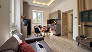 A modern living room with a sofa, a dining area with chairs, a kitchen with cabinets, and a TV mounted on the wall.