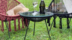 A glass table with a bottle and glass, surrounded by macramé chairs in a grassy outdoor area.