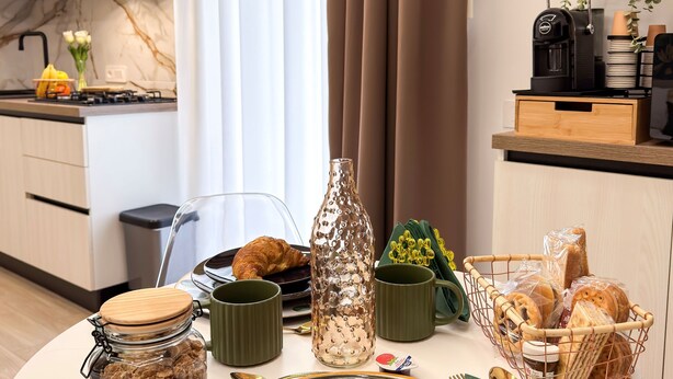 A modern kitchen with a dining table set for breakfast, including croissants, a glass jar, and a coffee maker.