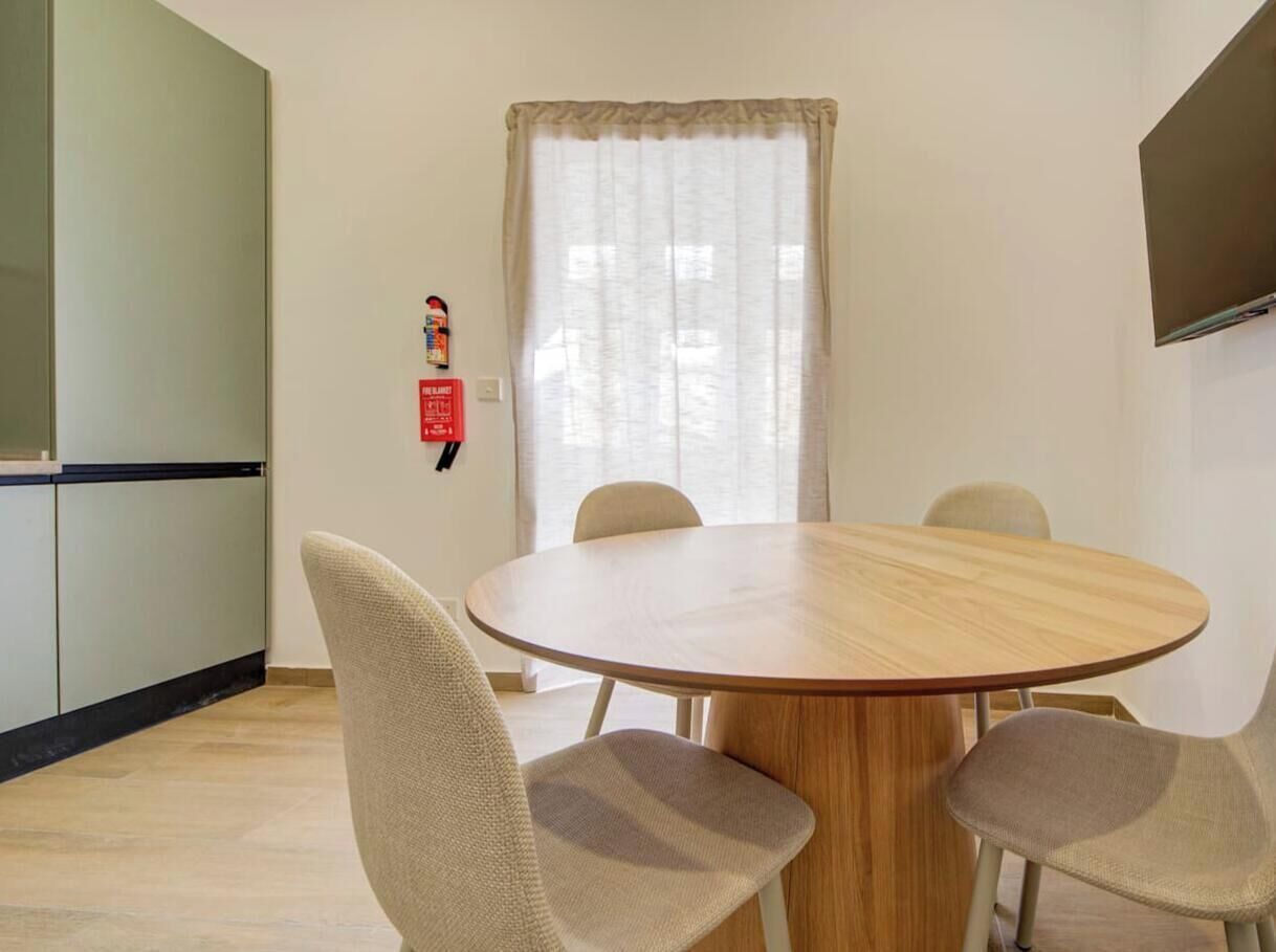 Un coin repas avec une table ronde en bois, quatre chaises, un extincteur fixé au mur et une fenêtre avec des rideaux.
