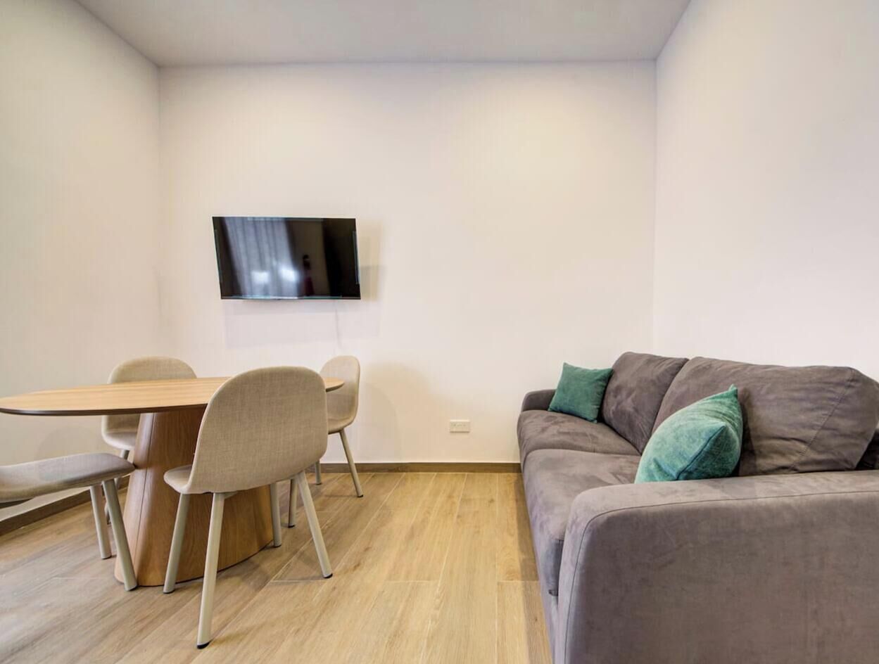 Une pièce comprenant une table à manger en bois, quatre chaises, un canapé gris avec des coussins verts et un téléviseur à écran plat fixé au mur.