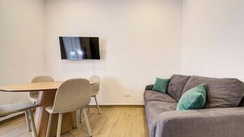 Une pièce comprenant une table à manger en bois, quatre chaises, un canapé gris avec des coussins verts et un téléviseur à écran plat fixé au mur.