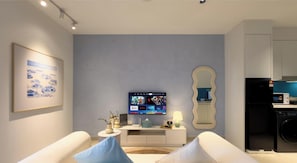 A modern living room with a flat-screen TV, a white sofa, a decorative mirror, and a kitchen area with appliances. - The Shore Kota Kinabalu Borneo Grand Roam Suites (Kota Kinabalu)