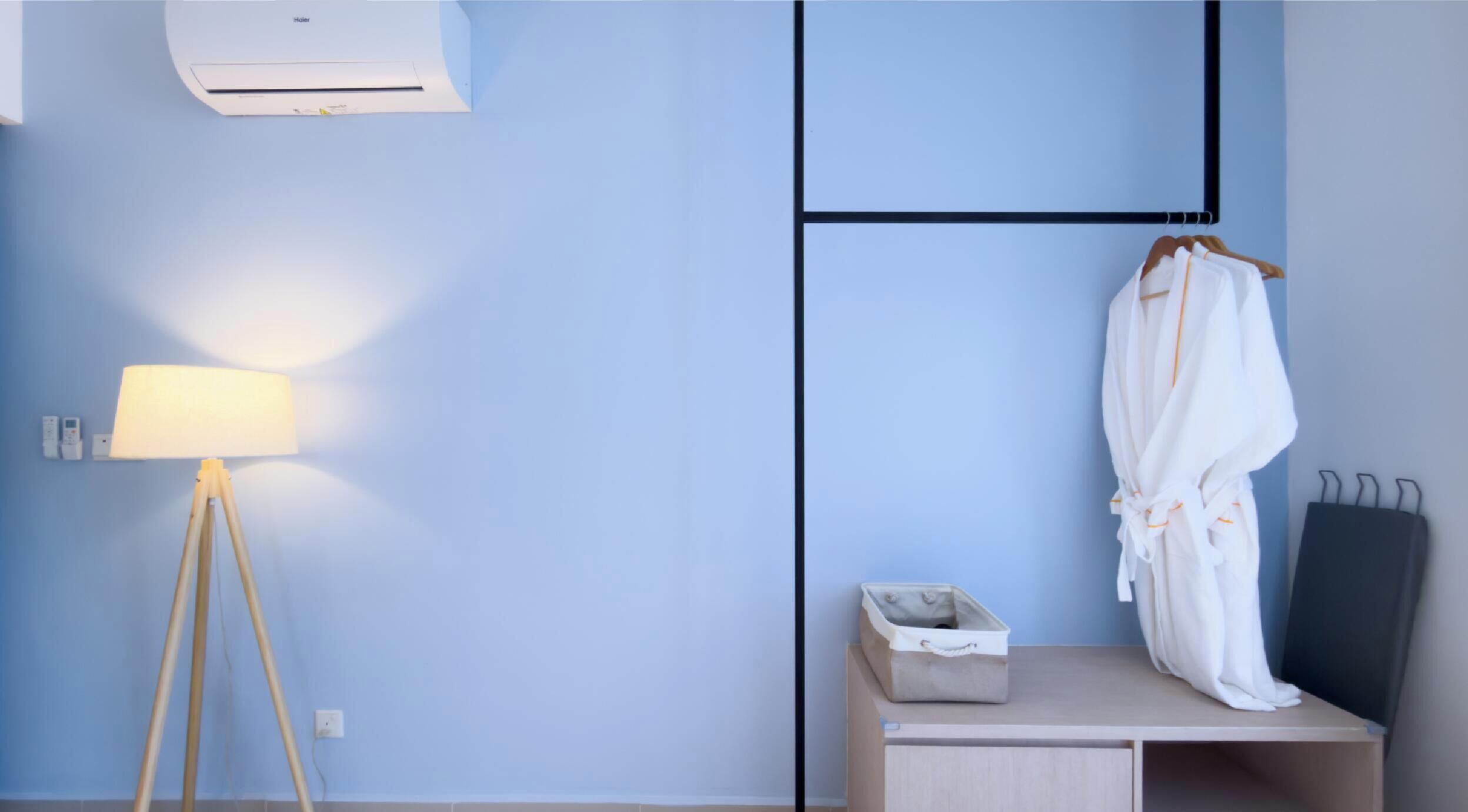 A room with a lamp, a robe hanging on a rack, a bedside table with a small box, and a wall-mounted air conditioner.