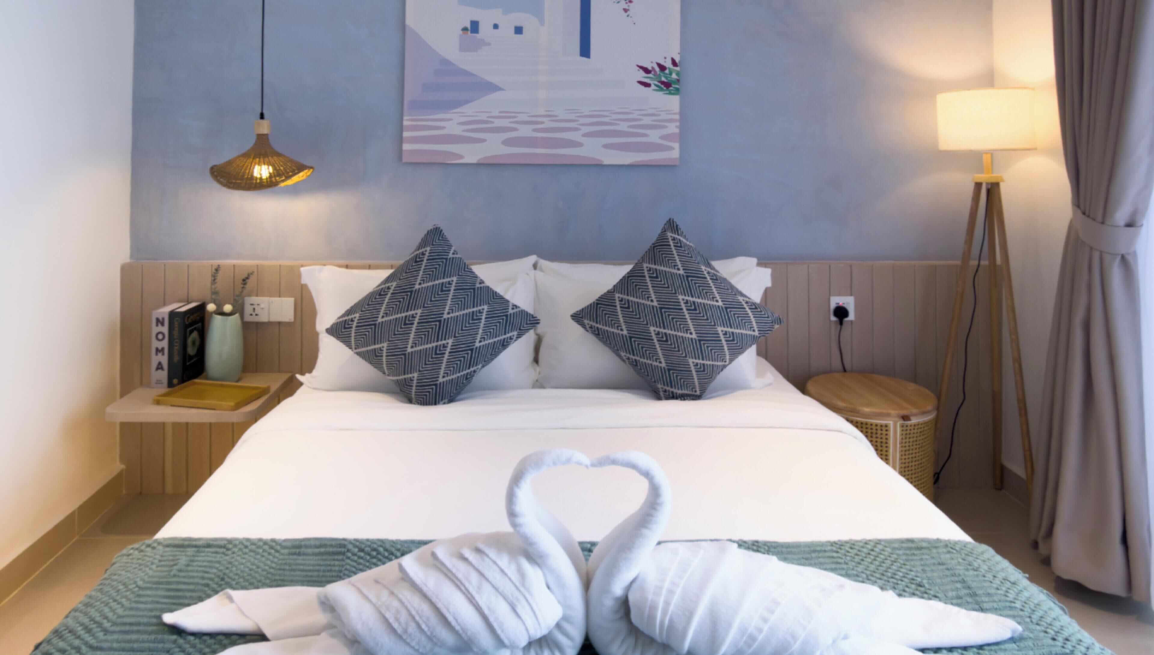 A neatly made bed with two pillows, a bedside lamp, a small table with a book, and a wall painting.