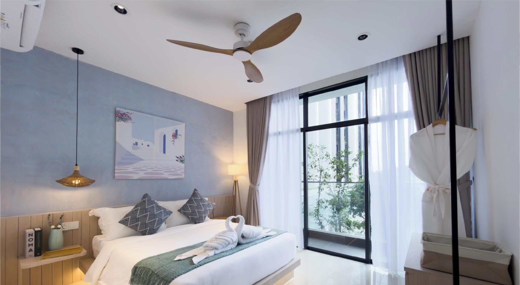 Ein Hotelzimmer mit Bett, Deckenventilator, großem Fenster mit Vorhängen, einem Gemälde an der Wand und einer Nachttischlampe.