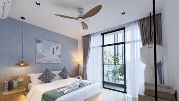 Ein Hotelzimmer mit Bett, Deckenventilator, großem Fenster mit Vorhängen, einem Gemälde an der Wand und einer Nachttischlampe.