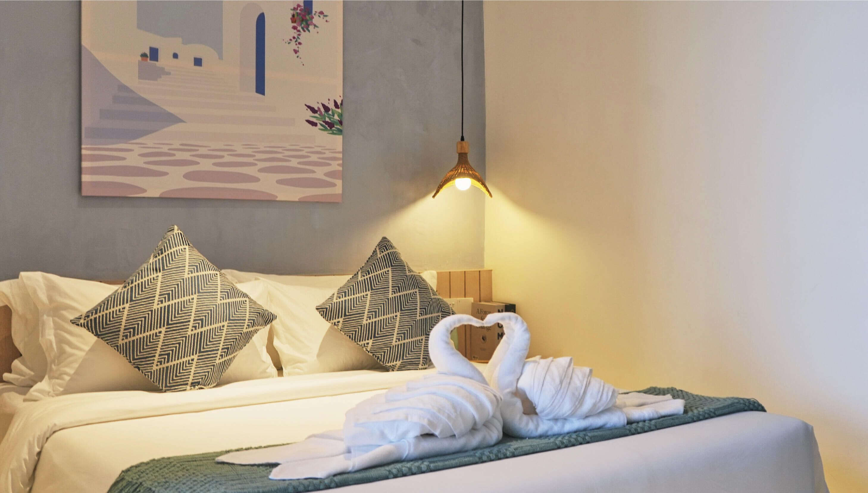 A hotel room with a bed, pillows, a wall-mounted lamp, and a painting.