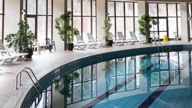Una piscina interior con suelo de baldosas, sillas de descanso y ventanas amplias.