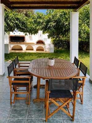 A covered patio with a round wooden table and chairs, an outdoor grill, and a white wall with arches.