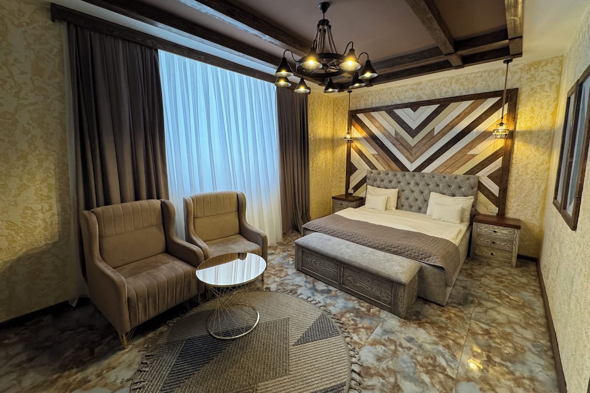 A hotel room with a bed, two armchairs, a small round table, a patterned rug, and a decorative wall with a geometric design.