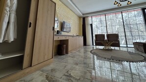 A hotel room with a wardrobe, a desk with a flat-screen TV, a large window with a view of trees, and a chandelier. - Tengealti Resort Hotel (Quba)